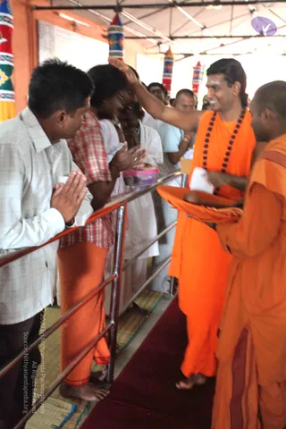 A photograph of a Sarva Darsan style gathering under a tent with striped and patterned banners. A man in long swept-back hair and an orange robe with a dark rudraksha mala raises his right hand in blessing toward a devotee. Beside him, a monk in orange holds an orange-cloth tray with a small object on it. On the left, a man in a white checked shirt stands with palms pressed together in namaskar, and a man in a red-and-white checked shirt with an orange lower cloth stands with hands folded. Behind them, a person in white clothing holds a small pink container. A red carpet runs along the aisle and a metal railing separates the crowd from the walkway.