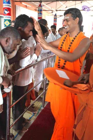 A photograph at a Sarva Darshan style gathering in a covered hall with striped red, blue and yellow pillars. A man in an orange robe with a dark rudraksha mala and a white tilak on his forehead raises his right hand in blessing over the head of a devotee in a white shirt, who bows with palms pressed together. Beside them, a man in a white shirt and an elderly man with folded hands also bow. An attendant in orange holds an orange tray with a folded cloth. Other people stand in the background against a metal railing, and a red carpet covers the floor. The orange-robed blessing figure has long swept-back hair and ornate ceremonial appearance, consistent with the page's a person reference, though the image does not confirm identity beyond appearance.