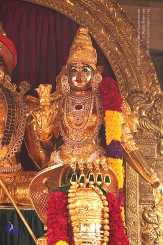 A golden deity murti in a procession or temple setting, seated with a tall gold crown and layered jewelled necklaces with red, green and white stones. The figure holds a raised gold object in one hand and wears a gold-framed arch behind. Beside it at left is a second gold figure in a red turban and ornate necklaces. Red and yellow-purple flower garlands drape over the central figure and a gold chariot-like base with green gem-set fittings sits in front. Red curtains and ornately carved gold panels form the backdrop. No living people are visible.