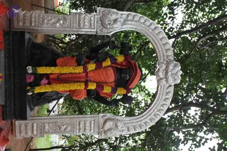 A dark stone statue of a seated saint in an ornate silver arched frame, set outdoors among green leaves. The figure wears an orange-saffron cloth draped over the body with a maroon edge, a rudraksha mala across the chest, and bold garlands of yellow marigolds interspersed with purple and pink flowers. Its hands are raised in a gesture beside the arch. The silver frame has carved lion-like motifs and foliage. The statue is a murti, not a living person, so no physical people are present.