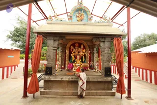 A photograph of an outdoor shrine under an open tin-and-red-pipe canopy. A stone shrine with carved pillars and a golden arched doorway holds a deity murti of a seated pair with a colourful painted backdrop, flanked by brass lamps and a garland of yellow flowers. Orange curtains hang at the sides. A single person in white dhoti and shawl, with a bare upper body, stands at the base of the shrine offering or worshipping before the murti, a puja scene. Trees and orange-and-white painted walls surround the open courtyard.