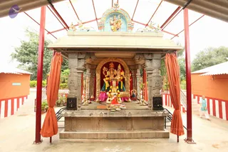 A stone shrine with carved pillars and a gold-framed arched niche stands under an open orange-and-red roofed canopy supported by red metal posts. Inside the niche, a pair of deity murtis are decorated with garlands, a red cloth backdrop behind them, and brass oil lamps flank the platform. Offerings and a small plate sit at the base. Orange curtains hang at the sides, and a painted panel with divine figures crowns the top. A woman in a light blue and white outfit walks past on the right. White walls with red and white bands frame the courtyard, and greenery is visible behind.