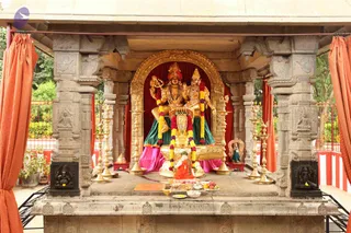 A photograph of an outdoor temple shrine with carved grey stone pillars and an open roof. Inside a golden arch framed by red velvet backdrop stands a gilded deity murti pair, one figure wearing a red and gold crown and holding a trident, the other in a golden crown beside it. Both wear green and pink skirts and garlands of yellow and magenta flowers. Brass oil lamps stand on either side, and a flower-garlanded object sits on the altar. Below, a small figure in orange clothing stands in front of a tray of fruit, sweets and offerings. Orange curtains hang at the sides and green foliage shows beyond the railings. No living people are visible apart from the small figure in orange, which is a statue-like form and not clearly a physical person.