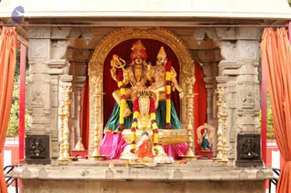 A golden shrine housing a pair of gilded deity murtis seated together on a raised golden throne. The figure on the left wears a red and gold turban, holds a trident-like staff, and wears rudraksha-style beads; the figure on the right wears a gold crown and ornate gold jewellery. Both are draped in green and pink silk, with a red velvet backdrop. Garlands of yellow and purple flowers hang from their shoulders. Golden oil lamps line both sides, and fruit and offerings sit before the throne. Orange curtains frame the sides of the shrine, and carved grey stone pillars surround the niche. No people are visible.