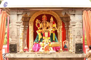 A golden deity murti of a standing and seated Shiva-Parvati pair under an arched gilded frame inside an open stone shrine pavilion. The male figure wears a red and orange turban, holds a trident, and is garlanded with marigolds in red and purple; the female figure wears a crown and ornate jewellery. Both sit on a golden throne-like peetha with pink and green draped cloth beneath. Brass oil lamps flank the shrine, orange curtains hang at the sides, and carved stone pillars frame the scene. A small figure in orange sits at the base, and a man in pale clothing with folded hands stands at the right edge, offering prayer.