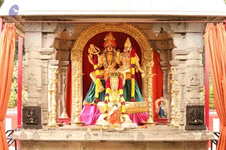 A photograph of an ornate golden shrine with gilded arch and carved stone pillars framing a pair of gilded deity murtis seated together on a golden throne. The figure on the left holds a trident and wears a red turban and jeweled necklace; the figure on the right wears a golden crown and red-and-gold garland. Both are draped in green and pink fabric. Yellow and red marigold garlands hang across the throne, and tall brass oil lamps stand on both sides. Orange curtains hang at the sides of the shrine, and a small seated figure in orange clothing sits at the base in front of offerings.