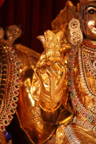 Close-up photograph of a gilded deity murti heavily adorned with jewelled ornaments. A large golden hand raised in a mudra holds a ornate golden staff or pillar with carved foliate motifs. Red, green and white gemstone necklaces and a decorated jewelled armlet cover the figure, set against a deep red curtain backdrop. The gold surfaces catch warm light. No physical people are visible; the figure is a statue.