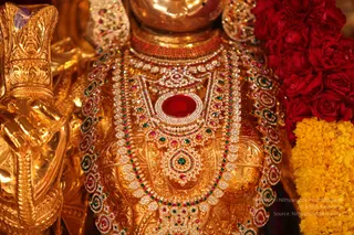 Close-up photograph of a gold deity murti adorned with heavy jewelled ornaments. The chest is covered by layered necklaces set with red, green and white stones, a large central red gem in a white-stone frame, and a dense band of pearls. Red roses and yellow marigold garlands hang at the right edge, and a gilded hand with a raised mudra is visible at the left.