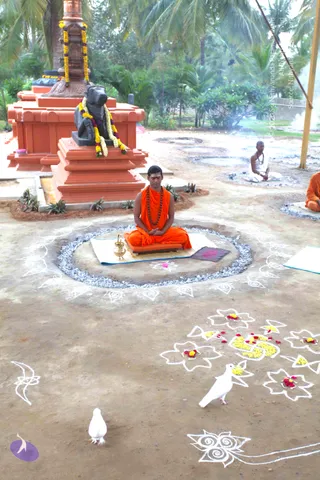 Outdoor photo of a seated ritual or meditation scene in a palm-fringed courtyard. A young man in orange saffron robes and a rudraksha mala sits cross-legged on a raised circular platform ringed with grey ash, with a brass kalasha-style vessel and a small cloth before him. Behind him stands an orange-and-red stepped shrine holding a black Nandi bull statue draped with yellow and red marigold garlands and a tall copper pillar. Another man in white dhoti sits cross-legged further back on a mat, and an orange-robed person sits at the right edge. White rangoli kolam designs with yellow and red flower petals decorate the ground, and two white doves are visible in the foreground.