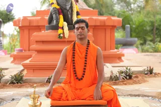 A man sits cross-legged in meditation on an orange cushion and mat outdoors, eyes closed, hands resting on his knees. He wears an orange sleeveless robe with a draped orange wrap, a long strand of dark rudraksha beads, and a red tilak on his forehead. Behind him stands a black stone deity statue draped with yellow, white and purple flower garlands, set on an orange tiered pedestal with a black Shivalinga to the right. A brass kalasha-style vessel sits at his left. Green plants and trees fill the background.