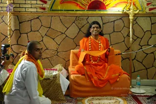 A photograph of a seated figure on an orange-cushioned throne in front of a stone-patterned wall with a gold sun-motif panel and gold trident staff behind. The seated man wears orange robes with gold trim, a rudraksha mala necklace, and a red bindi, with long dark hair swept back, smiling and holding an orange shawl. Beside him, a shaven-headed man in white kurta with an orange and yellow shawl kneels, holding a silver tray of flowers, while a camera operator with a flash unit is at the left edge. A water bottle and a patterned rug lie on the floor.