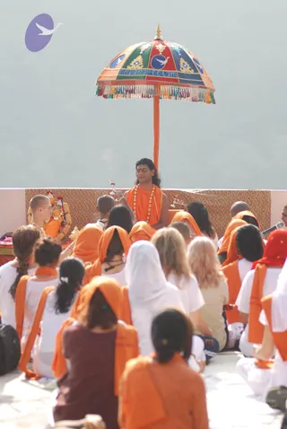 Outdoor satsang on a raised terrace under a hazy sky. A central seated figure in orange robes with a long orange-and-gold garland, red tilak and long swept-back hair holds a small ritual vessel while an umbrella canopy with the text "Dhyanapeetam" and a dove emblem shades him. Beside him, a deity murti decorated with marigold garlands and a priest in orange sits at a brocade-covered dais. Around them, a large seated gathering wears white and orange: many women in orange head coverings and orange sashes, some in white, a few in red shawls, and a man in orange. A microphone stand is at the right edge.