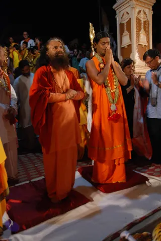 Night photograph of an outdoor riverside ceremony with a crowd behind. In the foreground, a bearded man in orange robes and a red shawl stands with palms pressed together in prayer, a rudraksha mala around his neck. Beside him, a younger man in saffron-orange robes with a garland of flowers and a gold-tinted ceremonial necklace stands with hands folded in anjali, eyes closed. Both stand on red cushions over white cloth. Behind them, a woman in black and a man in a white shirt with a camera also hold their hands together. A cream-coloured ornate stone pillar with carved bells rises at right, and people in yellow and orange garments fill the background.