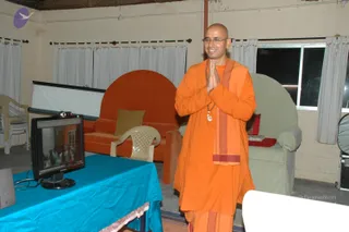A photograph of a bald man in orange saffron robes standing in a plain hall with cream walls and white curtains, palms pressed together in anjali namaskar with a smile. He wears round glasses, a rudraksha mala and a pendant, with an orange shawl draped over his shoulder. Behind him stands an orange sofa and a green armchair, and in front a computer monitor on a turquoise tablecloth shows a video feed of a crowd. A white plastic chair sits nearby.