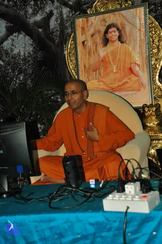 A seated man in orange robes and rudraksha mala sits in a cream armchair before a blue-draped table with a monitor, microphone and power strip, one hand raised to his chest. Behind him a framed portrait labelled with a copyright line shows a smiling figure with long hair, orange garments, a garland and a raised hand, set in a gilded frame beside a gold lion-form armrest and a painted tree backdrop with palm fronds.