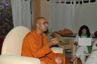 A photo of an indoor session in a plain room with white curtains and a folding table. A bald man in an orange saffron robe with a rudraksha mala sits in a cream upholstered chair, gesturing with his right hand while speaking. Beside him, a woman with curly dark hair in a white top, white trousers and a green scarf sits on a plastic chair, working on an open white laptop. A water bottle and an orange cloth rest on the table behind.