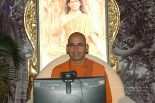 A bald man with glasses, wearing an orange saffron robe and a rudraksha mala, sits smiling in a white cushioned throne-style chair behind a computer monitor and webcam. Behind him, a gilded frame holds a large printed portrait of a smiling figure in orange robes with long dark hair, a rudraksha mala and a gold necklace, seated before a banyan-tree backdrop. A potted palm stands at left. The setting suggests an online satsang session.