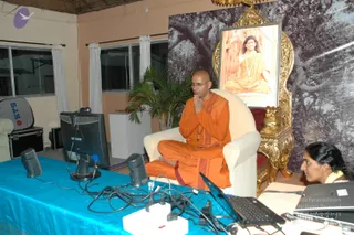 A seated man in saffron-orange robes with a shaved head and glasses sits in a white armchair in a plain room, hands joined in namaskar. Behind him on a gold ornate throne-style frame hangs a large portrait of a smiling figure in orange robes with long hair seated before a tree, and a black-and-white tree photograph covers the wall. A blue-draped table in front holds a Samsung monitor, small speakers, cables and a laptop, and a woman in the lower right corner works at a laptop. A potted palm stands near the chair.