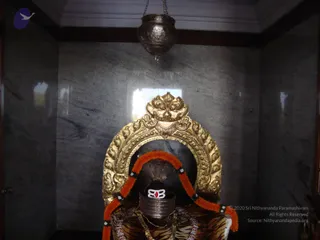 A photograph of a shrine niche with a grey polished stone wall and a hanging metal lamp. Centred is a deity murti seated before a large ornate gilded arch decorated with floral and foliate relief and a lion-face motif at the top. The murti wears an orange and white garland, a tiger-skin pattern cloth, gold necklaces and a silver-coloured headdress with a red stone ornament at the centre. No physical people are visible.