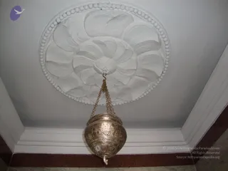 A photograph of a ceiling with a large white plaster rosette in the centre, shaped like a lotus with layered petals and a bead-border ring. Hanging from a hook at the centre is an ornate brass or silver-toned hanging lamp with embossed floral and scroll patterns on its bowl, suspended on fine chains. The walls are white with a dark red-brown dado along the floor edge. No people are visible.