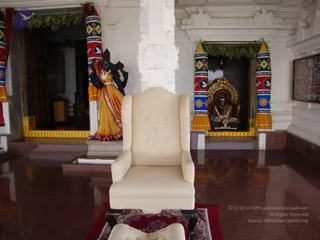 Empty cream upholstered wingback armchair with tufted back placed on a red and white patterned rug in a white temple hall with polished reddish floor. Behind it, a black stone deity murti in an orange-yellow sari stands in a niche decorated with striped red, green and white draped cloth with floral motifs and green leaf garlands. To the right, a shrine niche holds a gold-framed Shiva lingam with a rudraksha-style necklace and kumkum/vibhuti marking. A framed photo collage hangs on the right wall. No people are visible.