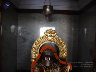A photograph of a shrine niche with a dark grey stone wall and a hanging silver lamp suspended from the ceiling. In front is a gold-framed arch with ornate floral and foliate carving and a gold crest at the top. Beneath it sits a dark shiva lingam with a white three-horizontal-line tripundra marking and a red bindi-like dot, wrapped in a tiger-striped cloth, with a metal yoni-like base and a garland of orange and white flowers draped over the arch. Gold necklaces hang below. No people are visible.