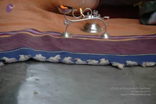 Close-up photo of a silver oil lamp with a lit flame and a small silver vessel on a stone floor. The lamp stands on a striped woven cloth with purple, blue, yellow and pink bands and cream tassels along the edge. Behind it an orange cloth and a copper or brass pot are partly visible. No people are visible in the frame.