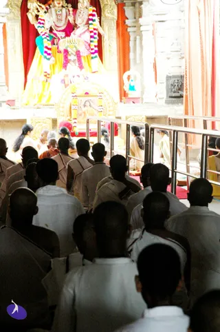 A photograph inside a temple hall during a morning worship service. Seated devotees in white and cream clothing, mostly seen from behind, fill the foreground and face a large decorated shrine. The shrine holds two deity murtis draped in multicoloured flower garlands, one with a red background and yellow cloth. Below them a framed picture garlanded with marigolds sits on a golden throne-like seat. In the background, seated figures in saffron and cream robes, including one in orange, sit near the shrine. Steel railings separate the seating areas, and a purple logo appears at the lower left corner.
