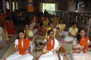 A photograph of a group of devotees seated on woven mats during a morning puja in a hall with red-brown walls and tall windows. In the foreground, several women in white dhotis or saris with orange shawls hold lit oil lamps (deepas) and small brass plates, some with palms pressed together. Behind them, many more devotees in white and yellow shawls sit in rows, a small boy in orange sits among them, and a yellow-and-orange hanging is visible on the upper wall. Brass bells and small offering plates with kumkum and flowers sit on the mats. Among the rows, the lamp flames and seated devotees fill the frame.