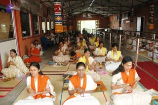 A large group of seated devotees performs a ritual in a covered hall with terracotta walls and a corrugated roof. In the foreground, women sit on woven mats with their hands folded in prayer, wearing white and cream saris with orange or yellow shawls. Several hold brass plates and small lamps, and brass puja vessels and bowls of flowers are arranged on the floor. Behind them, rows of white-clad participants sit on the mats beside a metal railing, and tall colourful decorative banners hang from the ceiling. No individual is identified as a person in this image.
