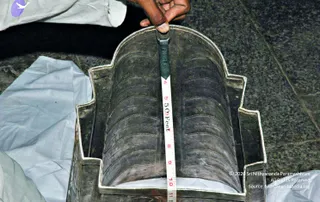 A close photograph of an ornate silver-coloured metal procession item. A person's hand, wearing a white sleeve, holds a green and white measuring tape against the object, marking its height in feet and inches. The object rests on a white cloth on a dark grey stone floor.