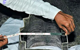 A close photo of hands measuring a dark grey, ribbed cylindrical object set in a metal frame or tray. A man in a white checked-sleeve shirt holds the object with his right hand while a second hand, at left, extends a white-and-red tape measure across it. The tape reads in feet and inches, with numbers visible around 4 to 12 and a marked 5 feet. The background is a dark stone or concrete floor with white cloth beneath the frame. No a person appears in this scene.