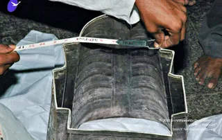 A photograph of a hand-held measuring tape being stretched across a dark, ridged metal object with a silvery rim. A man's hand holds the tape end in place while another hand, with bare feet visible at the lower right, rests on a white cloth. The grooved, cylindrical piece has a textured surface and a rectangular silver-edged base, and the scene is set on a dark floor covered with white cloth.