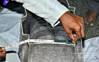A close photograph of a worker measuring an ornate silver procession item with a yellow-and-white retractable tape measure. A hand in a pale checked shirt holds the tape hook against the edge of a large silver-coloured base decorated with embossed scrollwork and a foliate pattern. A bare foot rests on a stone floor at the upper right, and a white cloth covers the ground beside the object. The tape reads in feet and inches and is held taut across the silver piece.
