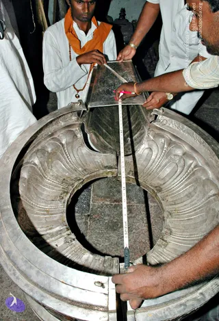 Photograph of a silver procession item being measured: a large circular silver-toned ornamental piece with repoussé lotus-petal or scalloped relief is laid out on a stone floor. Three men in white clothing handle the metal, one holding a silver plate with a red marker pencil while another measures with a tape. A young man in the centre wears an orange shawl, white kurta and a rudraksha mala, and stands at the left. A man in a checked shirt with a gold wristwatch works at the right edge. Four people are visible.