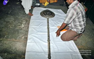 A photograph taken on a stone floor shows a long ornate silver ceremonial staff with a large heart-shaped top plate engraved with a floral medallion, lying on a white cloth. A man in a checked short-sleeved shirt and brown trousers kneels at the right, reaching toward the staff, while a barefoot person in white clothing stands at the upper left edge. A wooden board and a yellow cloth lie at the top of the frame.
