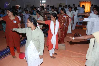 A photograph of a large indoor hall with an orange floor covering where many devotees stand in rows with arms extended forward, performing a synchronized standing exercise. In the foreground a woman in a sage-green cardigan over white clothing extends her arm, and beside her a woman in an orange kurta with a white shawl stands barefoot. Further back a woman in a red sari with gold border and a woman in red kurta with white trousers join the line. Many more people in white and pastel clothing fill the background. Name badges are visible on several chests, and a small red cushion lies on the floor at left.