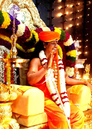 A seated man in saffron-orange robes and an orange turban sits on a gold-and-yellow throne (peetha) decorated with multicolour flower garlands of white, yellow, red and purple blossoms. He wears a long white and red flower garland, a layered gold necklace, rudraksha-style beaded bracelets and gold armlets, and smiles while raising one hand in greeting. Behind him are a golden deity-like backdrop, brass oil lamps and rows of small lit lamps on the wall. A probable Swamiji is the central figure, identified only by appearance cues: saffron robes, orange turban and ornate ceremonial adornments in a throne setting. One person is visible.