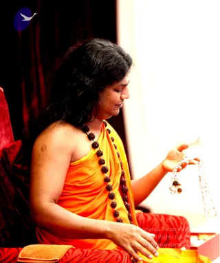 A photograph of a seated person in profile wearing a bright orange-yellow shawl draped over one shoulder, with a dark rudraksha mala worn across the chest. Long dark wavy hair falls past the shoulders. The person holds a white beaded mala in one raised hand while the other rests on a red patterned cushion, with a red velvet throne-like seat behind. A small orange towel and a red box are visible at the lower edge.