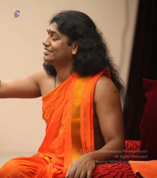 A photograph of a seated man in profile, eyes closed and smiling slightly, with long dark wavy hair falling past his shoulders. He wears an orange and gold-bordered wrap draped over one shoulder, leaving the shoulder bare, with a small red bindi on his forehead and a dark cord at his neck. His right arm is extended to the left of the frame. He sits on a red and black patterned cushion beside a deep red velvet pillow and dark drapery. The background is a plain beige wall.