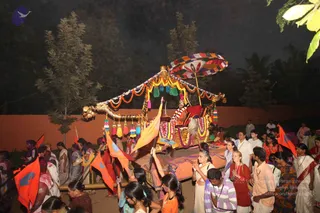 Night procession at an ashram: a decorated golden palanquin with a multicolour striped canopy and tasselled fringe, draped in garlands of marigolds and tinsel, carries a seated figure in red and white. Ahead and around it, devotees walk carrying orange and saffron flags. Many people in white, cream and saffron clothing, several women in saffron saris and rudraksha malas, follow along the orange wall. The seated figure has no clearly distinctive visible cues, so identity is uncertain.