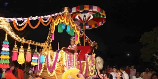 A night procession photographed from the side. A large golden palanquin-style vehicle with a multicolored canopy umbrella, hanging tassels in green, blue, yellow and pink, and bells is decorated with orange and purple marigold and white flower garlands. A seated figure in a red-and-gold crown-like headdress and a red garland sits on a red cushioned seat at the centre. Devotees in light clothing crowd around the vehicle, some reaching up to touch or hold it, and a figure at the lower right holds a white fan-like whisk. Tall palms and a dark sky with a moon frame the scene.