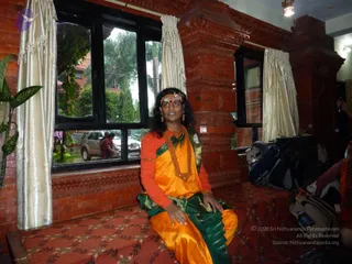A person seated on a red patterned carpeted bench inside a room with red brick walls and carved wooden pillars, beside tall windows with cream satin curtains. The person wears an orange blouse and an orange and green sari with gold trim, a long multi-strand orange-beaded necklace, a gold forehead band and a red bindi, and has long dark hair. Their hands rest on the bench as they look upward. Through the window a silver SUV and palm trees are visible outside, and bags and a water bottle sit on the floor to the right beside another person in orange. Appearance-based cues are not distinctive enough to identify anyone, so the person is described by role only.