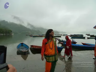 A photo taken on a misty lakeshore with rowboats moored in calm water and forested hills fading into fog. In the foreground a man with long dark hair stands smiling in an orange sari-style garment with a green border, a red blouse, a red beaded necklace and a forehead marking. A second figure in a red rain cape stands nearby, a boatman in red rain gear is visible in the background, and a person's hand holding a phone is at the left edge. The man in orange is a candidate for a person by appearance, but the cues are not distinctive enough to confirm, so the identity is unsure.