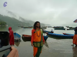 A woman with long dark hair and a red bindi stands on a wet lakeshore near a row of moored blue, red and wooden boats on misty water, with forested hills behind. She wears an orange sari with a green and gold border, matching orange blouse, a red beaded necklace and a red-and-white headband, and smiles toward the right. A person in a red raincoat stands at left, a boatman rows in the background, and a hand holding a phone is visible at left edge taking the photo. Another person in orange dhoti and a striped umbrella appear at right edge. Overcast, rainy scene.