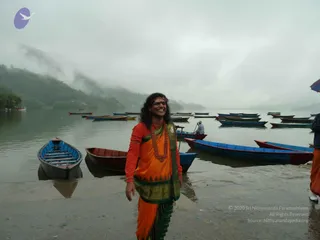 A smiling person stands on a wet stone landing beside a misty lake, with moored wooden boats in red, blue and green around them and forested hills fading into fog. The person wears an orange and red garment with a green-and-gold bordered wrap, a red beaded necklace and a red-orange headband, and holds the wrap at the side. A boatman in a white shirt sits in a small boat in the background, and a partially visible figure in orange stands at the right edge. The appearance is a long-haired figure in saffron-orange ceremonial dress, which is a person cue, but the page says he is not at this event and identity cannot be confirmed from appearance alone, so the value is unsure.