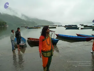 A photograph at a misty lakeshore with moored wooden boats, some red, blue and green, floating on grey water beneath wooded hills. In the centre, a figure with long dark hair wears an orange top with red sleeves, a bright orange and green wrap, and a long red-orange beaded necklace. Two young men in casual clothes stand at the water's edge to the left beside a blue rowboat. A partially visible person in white shoes appears at the right edge. a person candidate appearance is not clearly established from the visible cues, so this is recorded as unsure.
