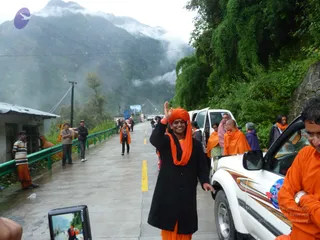 A photograph of a misty mountain road with a wet concrete surface and a green guardrail, lined with dense green forest. A man in an orange turban and orange shawl over a long black coat smiles and raises his right hand in a wave in the centre of the road. Around him, several people in orange and saffron robes stand beside a white SUV and walk along the roadside. A devotee with a camera lifts a small device in the lower left. Hills and cloud fill the background, with a roadside building at left.