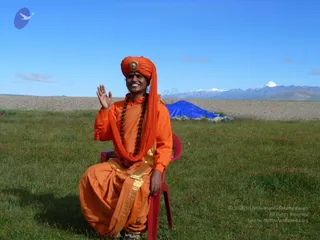 A smiling person sits on a red plastic chair on green grass, waving with the right hand. They wear an orange turban with a gold ornament at the front, an orange tunic with a gold-bordered orange wrap, and a long rudraksha mala. Behind them lies a dry gravel plain, a blue tarpaulin, and snow-capped mountain peaks under a clear blue sky.