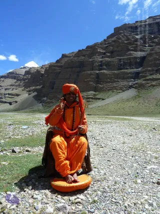 A smiling person in full saffron-orange robes and an orange turban with a gold ornament sits cross-legged on an orange cushion on a stony high-altitude plain. They raise one hand in a blessing gesture and hold a long trident-topped staff. Several rudraksha mala beads hang across the chest, and an orange shawl drapes over the shoulders above a leopard-print seat cover. Behind them rises a layered brown rock cliff under a bright blue sky, with a snow-capped peak visible at left. No a person candidate can be confirmed from appearance alone, so the person is described by role as a sannyasi-style figure.